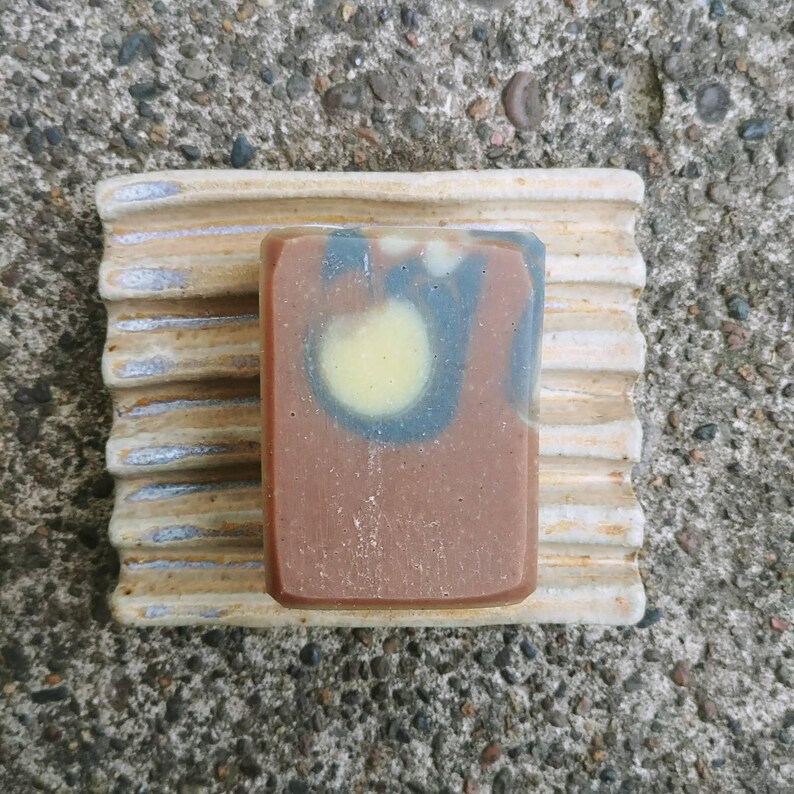 Puede incluir: Una pastilla de jab&oacute;n marr&oacute;n y negro con un dise&ntilde;o de remolino en un jabonera de cer&aacute;mica blanca.
