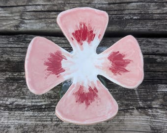 May include: A ceramic flower-shaped dish with a light pink exterior and a white center. The interior of each petal features a darker red brushstroke design. The dish is placed on a weathered wooden surface.