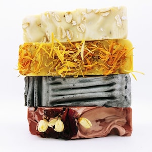 May include: Four bars of handmade soap stacked on top of each other. The top bar is white with oatmeal flakes, the second bar is yellow with dried flowers, the third bar is gray with a textured surface, and the bottom bar is pink with dried flowers.