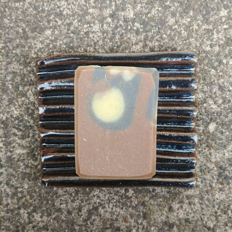Puede incluir: Una pastilla de jab&oacute;n marr&oacute;n y negro con un dise&ntilde;o de remolino se encuentra sobre un jabonera de cer&aacute;mica negra y marr&oacute;n.