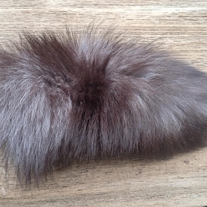 Könnte beinhalten: Ein flauschiges, ovales Fellstück mit einem Farbverlauf von Dunkelbraun zu Silbergrau. Das Fell ist dicht und wirkt weich, auf einer hellbraunen Holzoberfläche liegend. Die Textur des Fells wird durch das Licht hervorgehoben.