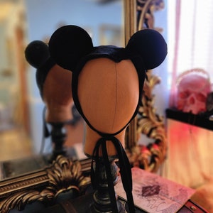 May include: A black felt Mickey Mouse hat with large ears. The hat is on a mannequin head and has a black ribbon tied around the neck.