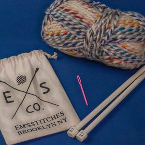 May include: A white drawstring bag with the text "E & S Co. EM'S STITCHES BROOKLYN NY" printed on it. The bag contains a ball of colorful yarn and two wooden knitting needles.