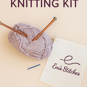 May include: A beginner knitting kit with a ball of lavender yarn, copper knitting needles, and a blue stitch marker. A white canvas bag with the text "Em's Stitches" is also included. The words "BEGINNER KNITTING KIT" are at the top.