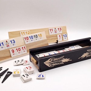 May include: A Rummikub game set featuring two light-colored wooden tile racks holding numbered tiles, a dark wooden storage box with decorative feather-like carvings, several stacks of white plastic tiles with colored numbers, and four black plastic stands.