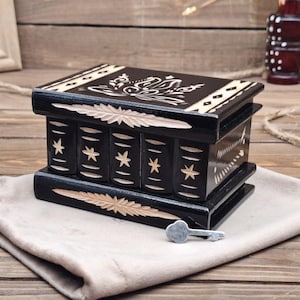 May include: A rectangular black wooden puzzle box featuring carved light-colored star patterns on the front and a floral design on the lid. The box has a horizontal carved leaf motif on the top and bottom edges and includes a small metal key.