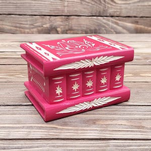 May include: A rectangular hot pink wooden box designed to resemble a stack of books. The box features white carved floral and star patterns on the top and sides, with horizontal carved leaf motifs along the top and bottom edges of the exterior.