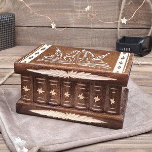 May include: A rectangular dark brown wooden box featuring a carved design on the lid and front. The front panel displays a series of vertical columns resembling book spines, each decorated with a small star. The lid has a floral pattern.
