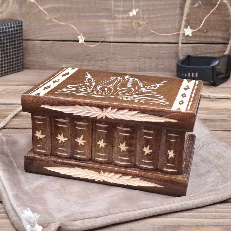 May include: A rectangular dark brown wooden box featuring a carved design on the lid and front. The front panel displays a series of vertical columns resembling book spines, each decorated with a small star. The lid has a floral pattern.