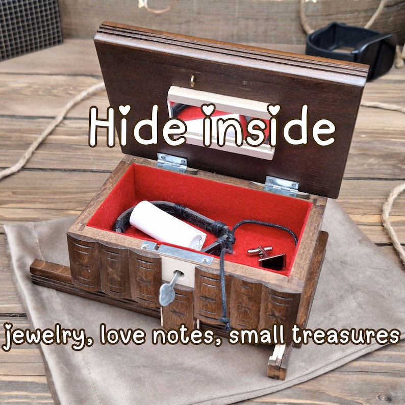 May include: A dark brown wooden puzzle box with a hinged lid featuring a mirror and the text Hide inside. The interior is lined with red fabric and contains a black bracelet, a white paper roll, and a cufflink. Text reads jewelry, love notes, small treasures.