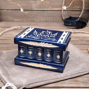 May include: A rectangular dark blue wooden puzzle box featuring light-colored carved floral patterns on the lid and star motifs on the front panel. The box has a smooth finish with horizontal carved leaf-like accents along the top and bottom edges.