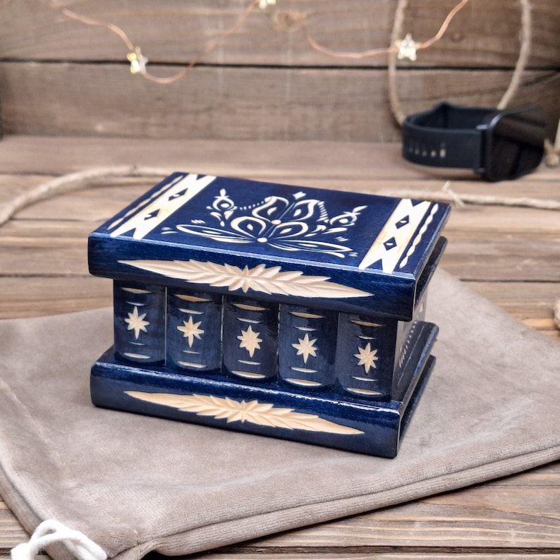 May include: A rectangular dark blue wooden puzzle box featuring light-colored carved floral patterns on the lid and star motifs on the front panel. The box has a smooth finish with horizontal carved leaf-like accents along the top and bottom edges.