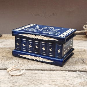 May include: A rectangular dark blue wooden box designed to resemble a stack of books. The lid and sides feature hand-carved light wood details, including star motifs, leaf patterns, and geometric lines. The surface has a smooth, polished finish.