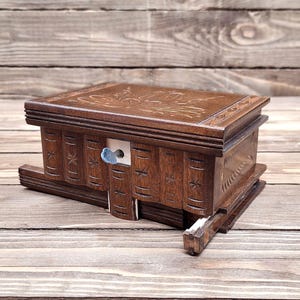 May include: A rectangular wooden puzzle box with a dark brown finish, featuring carved floral patterns on the lid and vertical grooves on the sides resembling book spines. A small blue metal key is inserted into the front panel of the box.