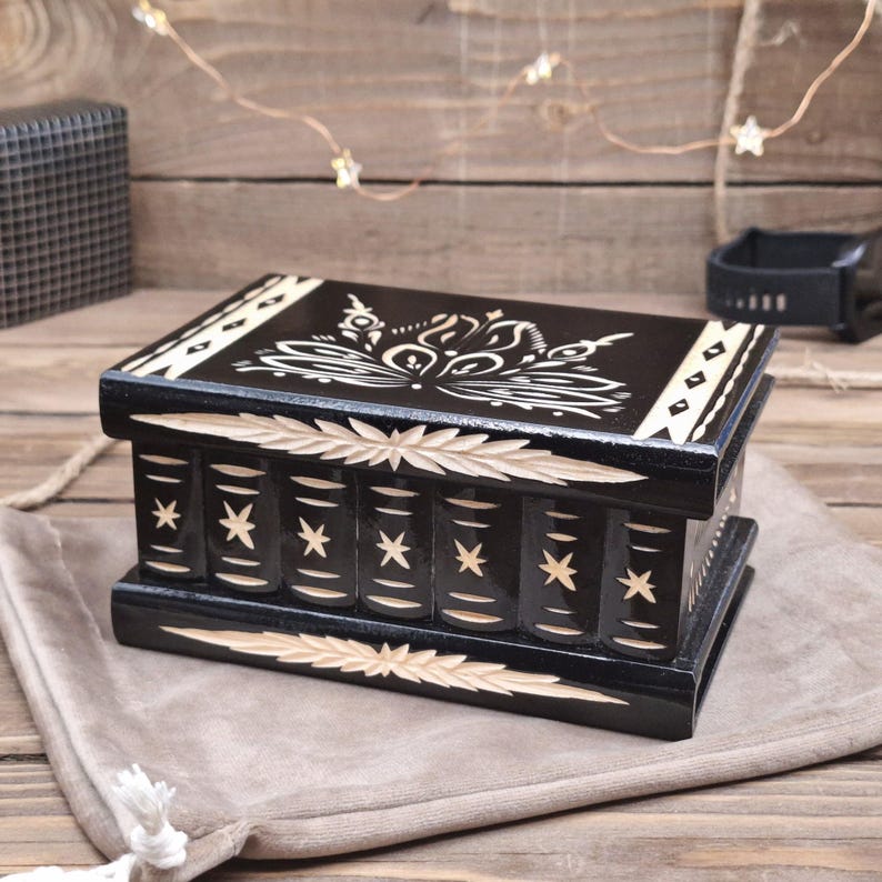 May include: A rectangular black wooden box with a hinged lid, featuring intricate light-colored carved floral patterns on the top and star motifs on the front panel. The box has a polished finish and decorative carved borders along the top and bottom edges.