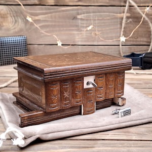May include: A rectangular dark brown wooden box with a carved top and a front panel designed to resemble a row of books. The box features a sliding mechanism with a small metal key protruding from the center, and a pair of silver cufflinks nearby.