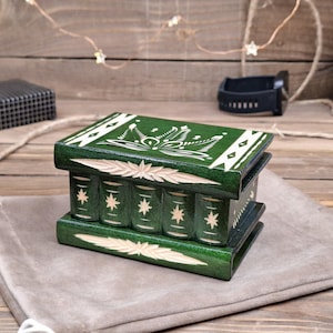 May include: A rectangular dark green wooden box designed to resemble a stack of books. The lid and base feature carved leaf patterns, while the sides display vertical book spine carvings with star motifs. The box has a smooth, polished finish.