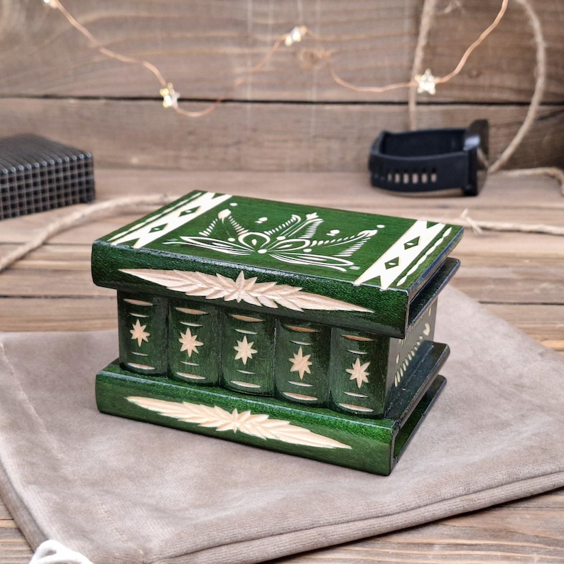 May include: A rectangular dark green wooden box designed to resemble a stack of books. The lid and base feature carved leaf patterns, while the sides display vertical book spine carvings with star motifs. The box has a smooth, polished finish.