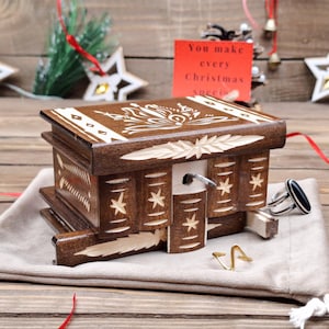 May include: A dark brown wooden puzzle box with white carved floral and star patterns on the lid and sides. The box features a sliding front panel and a small metal key. A silver ring with a black stone and a gold wire ring sit nearby.