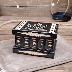 May include: A rectangular black wooden box with a hinged lid, featuring carved light-colored floral patterns on top and star motifs on the front panels. The box has a smooth, polished finish and is designed with a decorative border of diamonds.