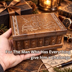 May include: A rectangular wooden puzzle box with a dark brown finish, featuring an intricate floral carving on the lid and a side design resembling a row of small books with star accents. The text reads: For The Man Who Has Everything ... give him a challenge.