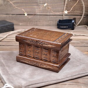 May include: A rectangular wooden box with a dark brown finish, featuring intricate hand-carved floral and star patterns on the lid and sides. The front panel is designed to resemble the spines of several small books, each decorated with a carved star.