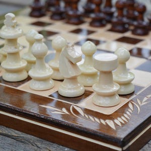 May include: A wooden chess set with light and dark pieces arranged on a checkered board. The light pieces are ivory-colored, and the dark pieces are brown. The board is framed in dark wood with decorative carvings.