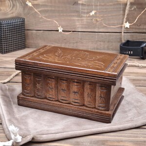 May include: A rectangular dark brown wooden box with a hinged lid featuring an engraved floral pattern. The front side of the box is carved to resemble the spines of several books arranged side by side, each decorated with a small star-shaped motif.