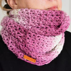 May include: A chunky knit infinity scarf featuring a gradient of pink, magenta, and white yarn with subtle metallic silver threads woven throughout. A small rectangular tan leather tag with the word Handmade is attached to the lower edge.