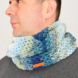 May include: A chunky crochet cowl scarf featuring a variegated yarn pattern in shades of blue, light blue, white, and pale green. The neck warmer has a textured, open-stitch design and includes a small, rectangular orange leather tag labelled Handmade.