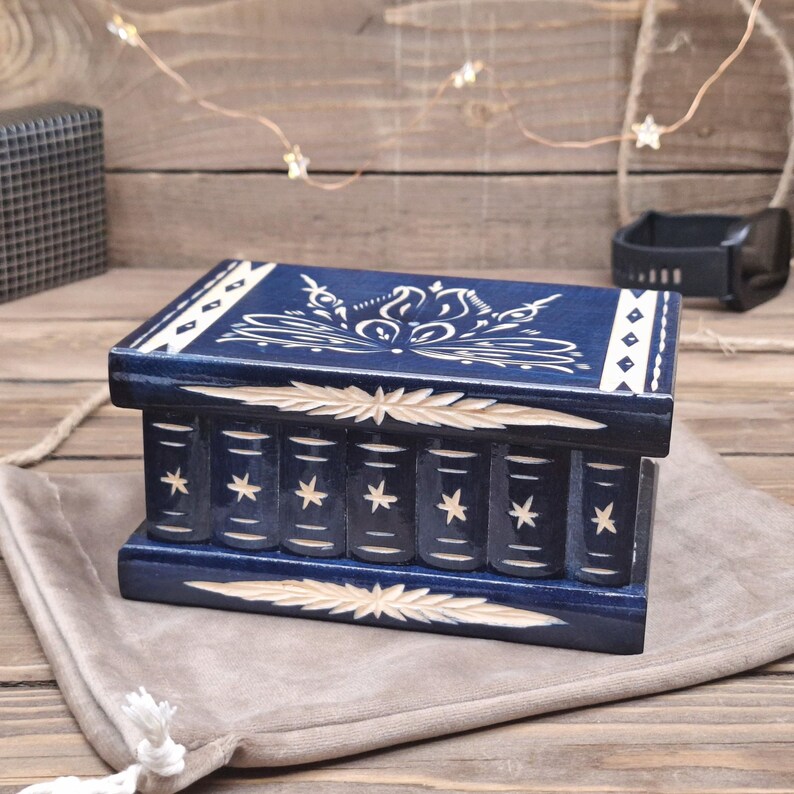 May include: A rectangular dark blue wooden puzzle box featuring carved white star motifs on the front panel and a decorative floral pattern on the lid. The box includes horizontal carved leaf-like accents on the top and bottom edges.