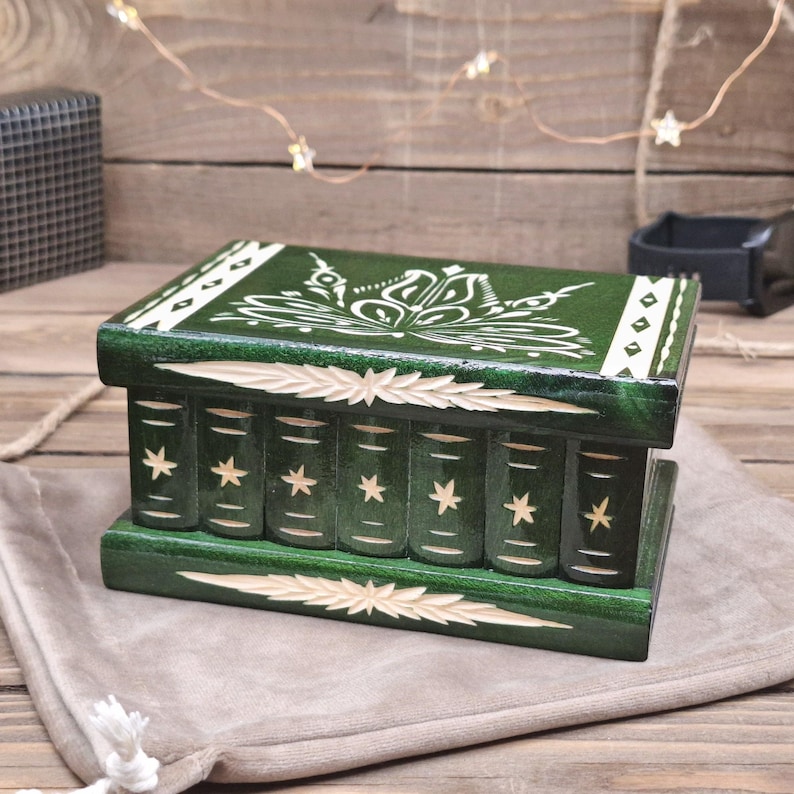 May include: A rectangular dark green wooden box with a hinged lid. The lid features a white floral design and geometric borders. The front panel is carved to resemble a row of seven book spines, each decorated with a small white star motif.