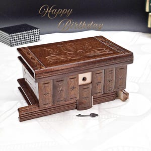 Op de afbeelding: Een donkerbruine houten puzzelbox met een bloemmotief op het deksel en sterrensnijwerk aan de zijkanten. Een kleine sleutel ligt bij de doos. De woorden "Happy Birthday" staan in gouden letters erboven.