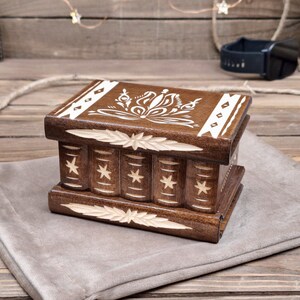 May include: A rectangular dark brown wooden puzzle box featuring a white floral design on the lid. The sides are carved with vertical columns, each decorated with a small star. The top and bottom edges have carved leaf-like patterns.