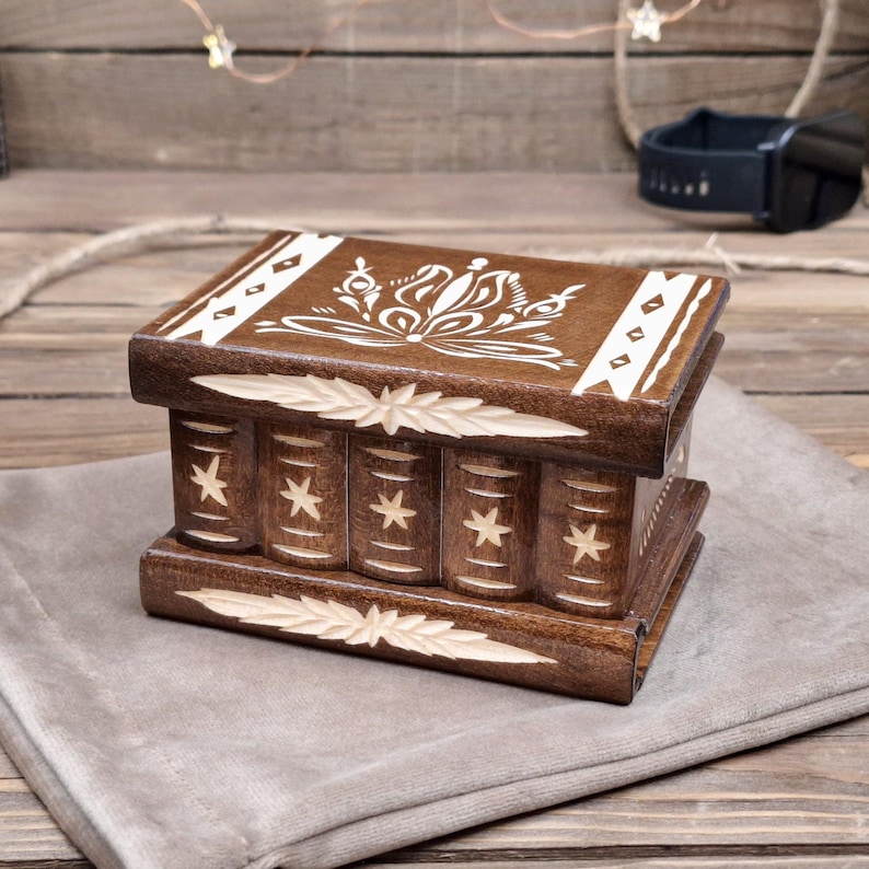 May include: A rectangular dark brown wooden puzzle box featuring a white floral design on the lid. The sides are carved with vertical columns, each decorated with a small star. The top and bottom edges have carved leaf-like patterns.