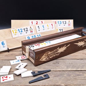 May include: A wooden Rummikub game set with a dark brown box and a light wood tile holder. The box is decorated with a feather design. The tiles are white with colored numbers. The game is displayed on a wooden surface.