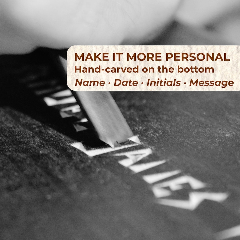 May include: A metal chisel carves the name JAMES into a dark, flat surface. A text overlay reads: MAKE IT MORE PERSONAL, Hand-carved on the bottom, Name &middot; Date &middot; Initials &middot; Message. The image is in black and white with a focus on the carving tool.