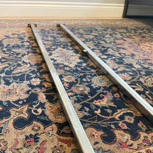 Peut inclure: Deux pieds de lit en métal argenté avec des supports carrés aux extrémités. Les pieds sont posés sur un tapis à motifs.