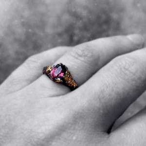 May include: A ring with a dark metal band and a large, oval-shaped, dark red gemstone. The ring is worn on a person's finger.
