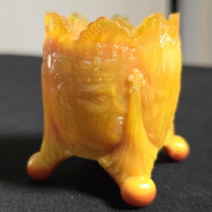 May include: A vintage, amber-colored glass egg cup with a detailed face design. The cup has three rounded feet and a scalloped rim. The glass has a glossy finish, and the overall design is ornate and decorative.