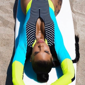 Puede incluir: Una mujer con un traje de baño de rayas azules, amarillas y negras de manga larga. Está tumbada sobre una tabla de surf blanca con el pelo recogido en una coleta.
