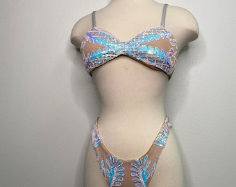 May include: A two-piece swimsuit with a sheer, nude fabric base and iridescent blue and purple sequins. The top is a bandeau style with thin straps and the bottom is a high-cut style.