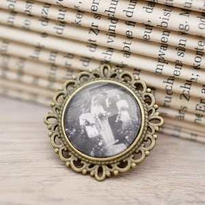 Könnte beinhalten: Eine Brosche im Vintage-Stil mit einem goldfarbenen Metallrahmen und einem Glas-Cameo mit einem Schwarzweißfoto einer Hochzeitsgesellschaft.