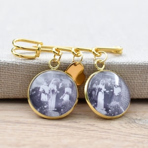 Könnte beinhalten: Goldfarbene Brosche mit zwei runden Fotoanhängern. Jeder Anhänger zeigt ein Schwarzweißfoto einer Hochzeitsgesellschaft. Die Brosche hat einen herzförmigen Anhänger.