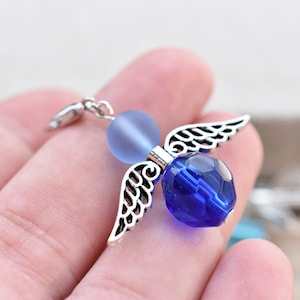 Può includere: Un ciondolo a forma di angelo in argento con perline di vetro blu. L'angelo ha ali delicate e una perla blu opaca per la testa.