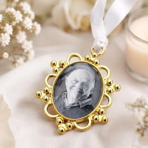 Può includere: Un ornamento commemorativo dorato con una foto ovale di una persona. L'ornamento ha una cornice decorativa ed è sospeso da un nastro bianco. Una candela accesa e fiori bianchi sono sullo sfondo.