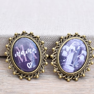 Könnte beinhalten: Zwei ovale Broschen mit verzierten goldenen Rahmen. Eine Brosche zeigt einen violetten Hintergrund mit dem Wort "mama" in rosa Schrift und einem kleinen weißen Herzen. Die andere Brosche zeigt ein Schwarzweißfoto einer Frau, die auf einem Friedhof steht.