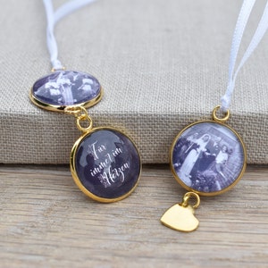 Könnte beinhalten: Zwei goldfarbene Anhänger mit weißen Satinbändern. Ein Anhänger zeigt ein Schwarzweißfoto einer Gruppe von Menschen, der andere Anhänger zeigt den Text "Für immer im Herzen".