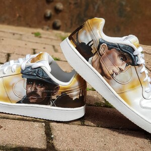 May include: A pair of white trainers with a custom design featuring a portrait of a man wearing a cap and a city skyline in the background. The trainers have a yellowed effect and a white swoosh logo.