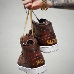 May include: A pair of brown high-top sneakers with white soles. The words "JUST" and "MARRIED" are printed in gold on the heels. The shoes are held by their laces.